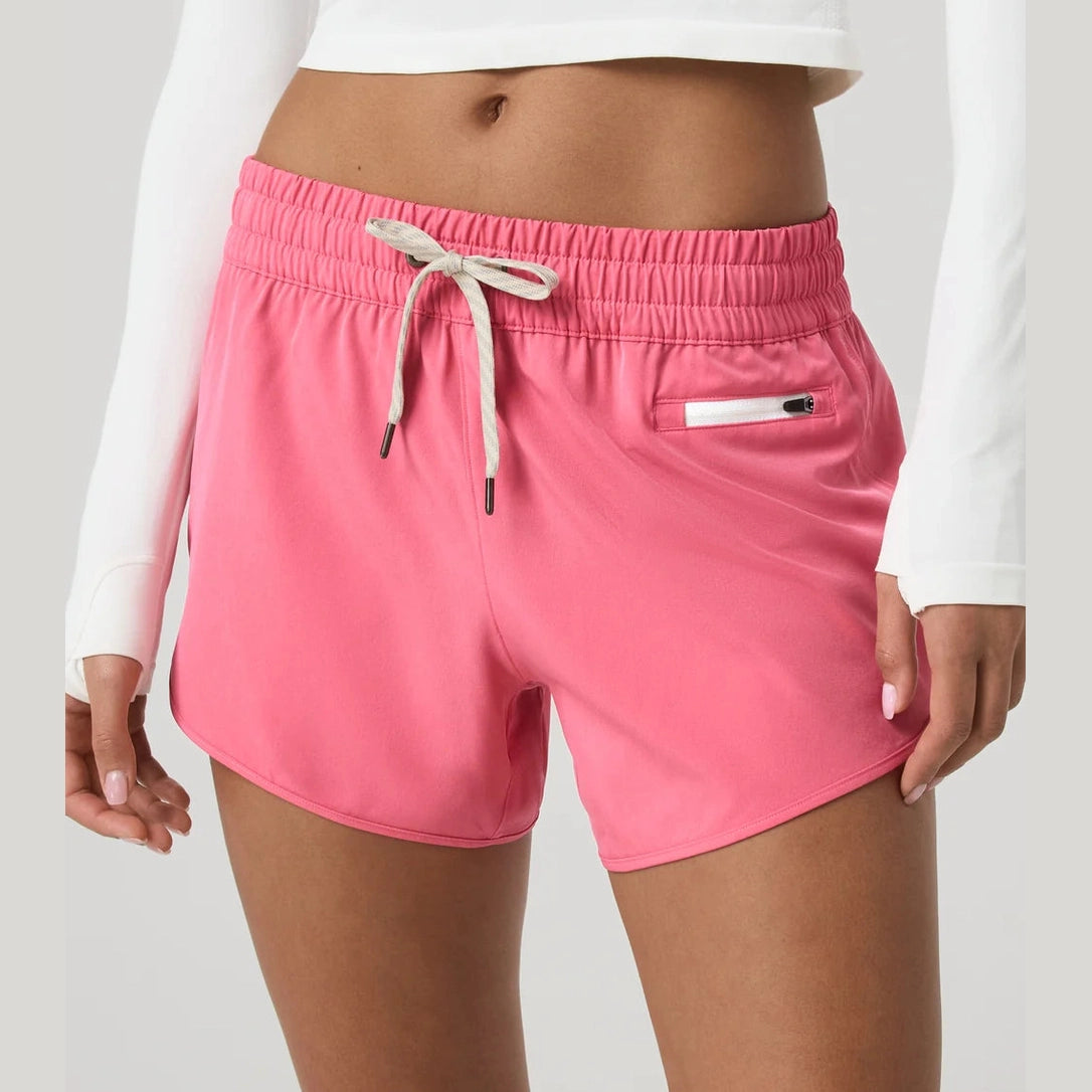 A model wearing bright pink Vuori Clementine 4" Short – Women's athletic shorts with a white drawstring and a zippered side pocket, paired with a white long-sleeve crop top against a light gray studio background.