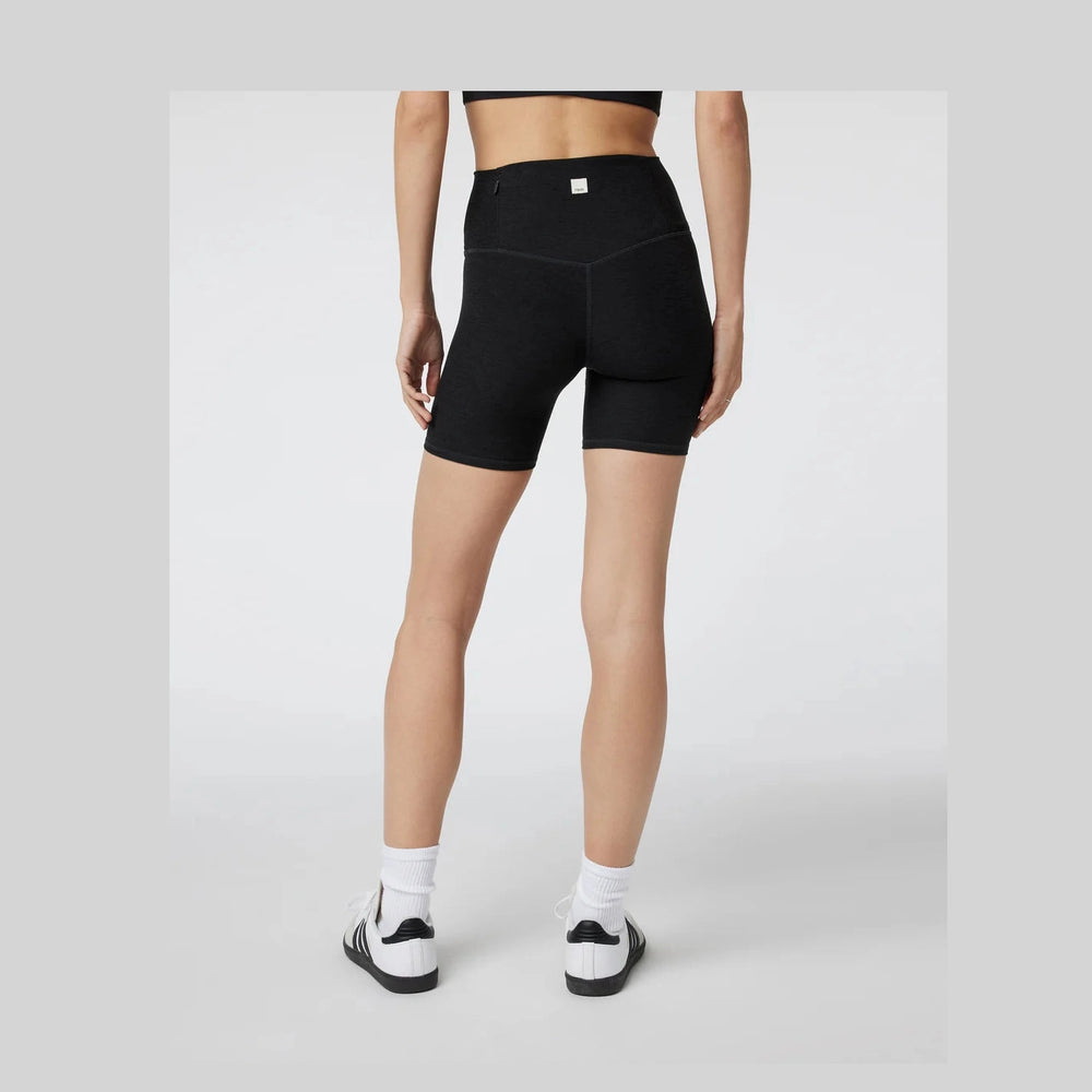 Rear view of a model wearing black Vuori Clean Elevation Shorty – Women's high-waisted biker shorts, white socks, and black-and-white sneakers against a light gray studio background.