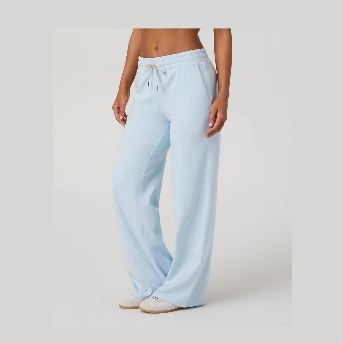 A model wearing light blue wide-leg Vuori Halo Essential Wideleg – Women's pants with an elastic drawstring waist, paired with white slip-on shoes, against a neutral light gray studio background.