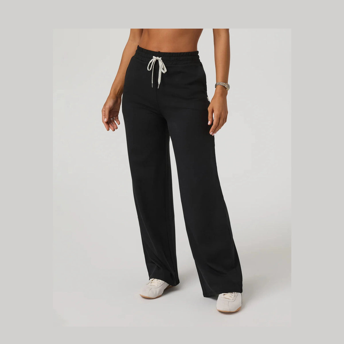 A model wearing black Vuori Halo Essential Wideleg – Women's wide-leg lounge pants with a white drawstring waist, paired with white sneakers, shown against a light gray studio background.