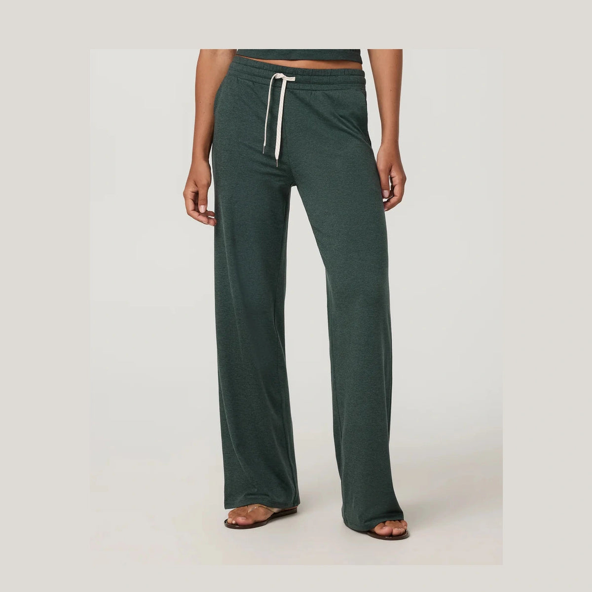 A person wearing dark green wide-leg Vuori Halo Essential Wideleg – Women's pants with a white drawstring, shown from the waist down against a light gray studio background.
