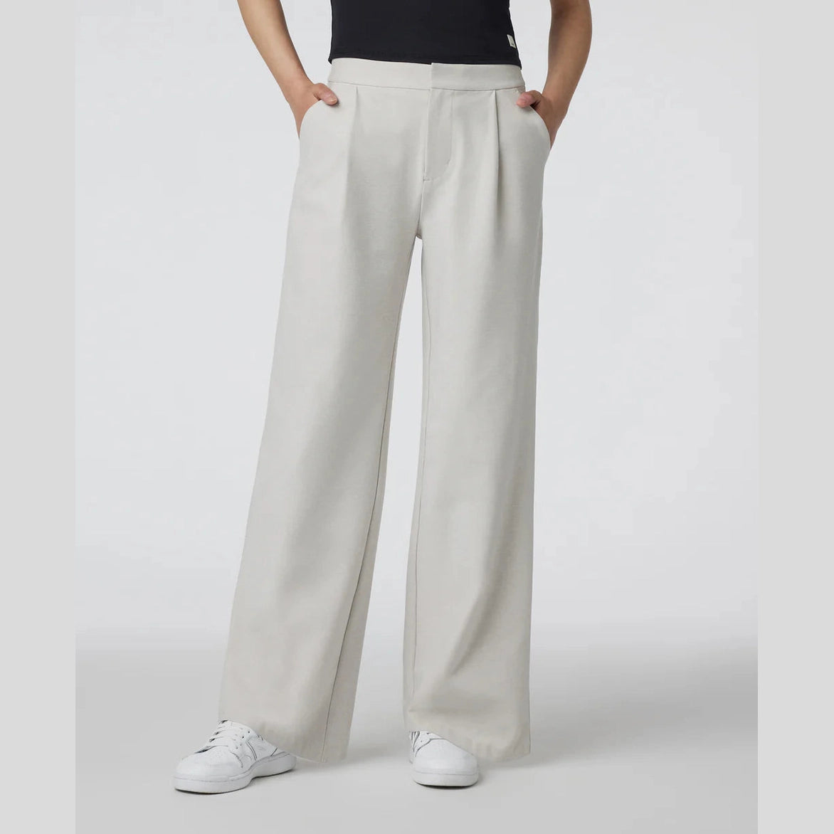 A model wearing Vuori Elevation Trouser – Women's high-waisted, wide-leg trousers in a light oatmeal beige color, paired with a black top and white sneakers, shown from the waist down against a light gray studio background.