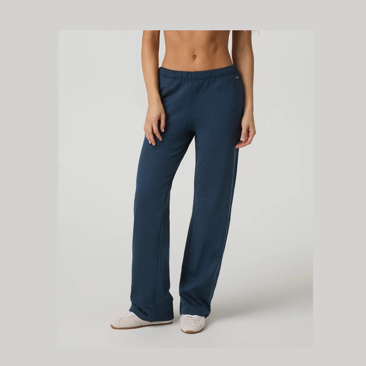 A model wearing dark blue wide-leg Vuori Halo Modern Straight Leg Pant – Women's pants and white slip-on shoes, shown from the torso down against a light gray studio background.