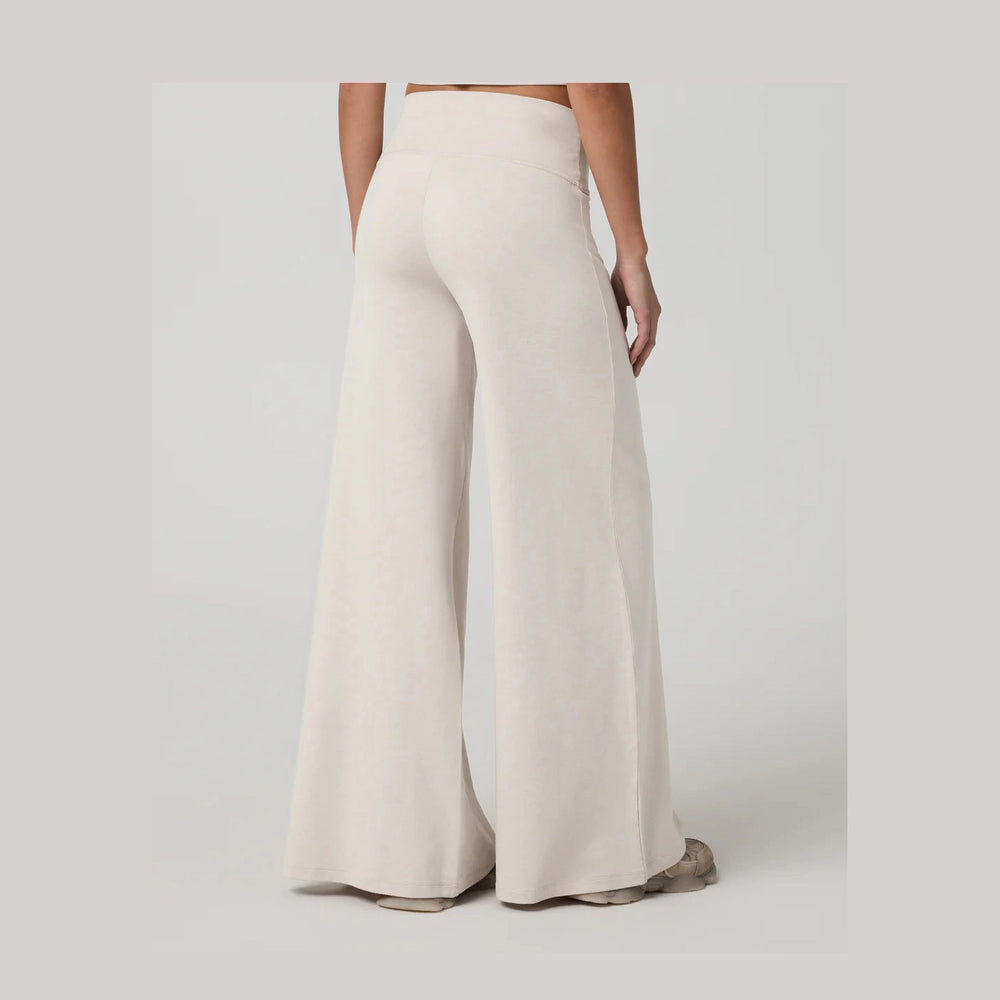 A person shown from the back wearing high-waisted, Vuori Halo Easy Wideleg Pant – Women's pants in a light cream or off-white color, paired with gray shoes, against a plain light gray studio background.