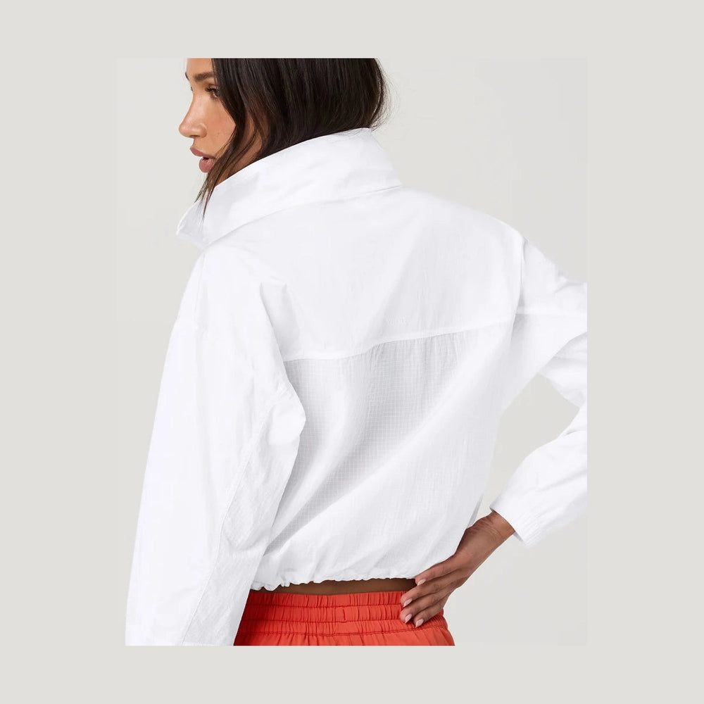 A close-up back view of a model wearing a white Court Full Zip--Women's jacket with a high collar and textured mesh paneling, paired with red pants, against a light gray studio background.