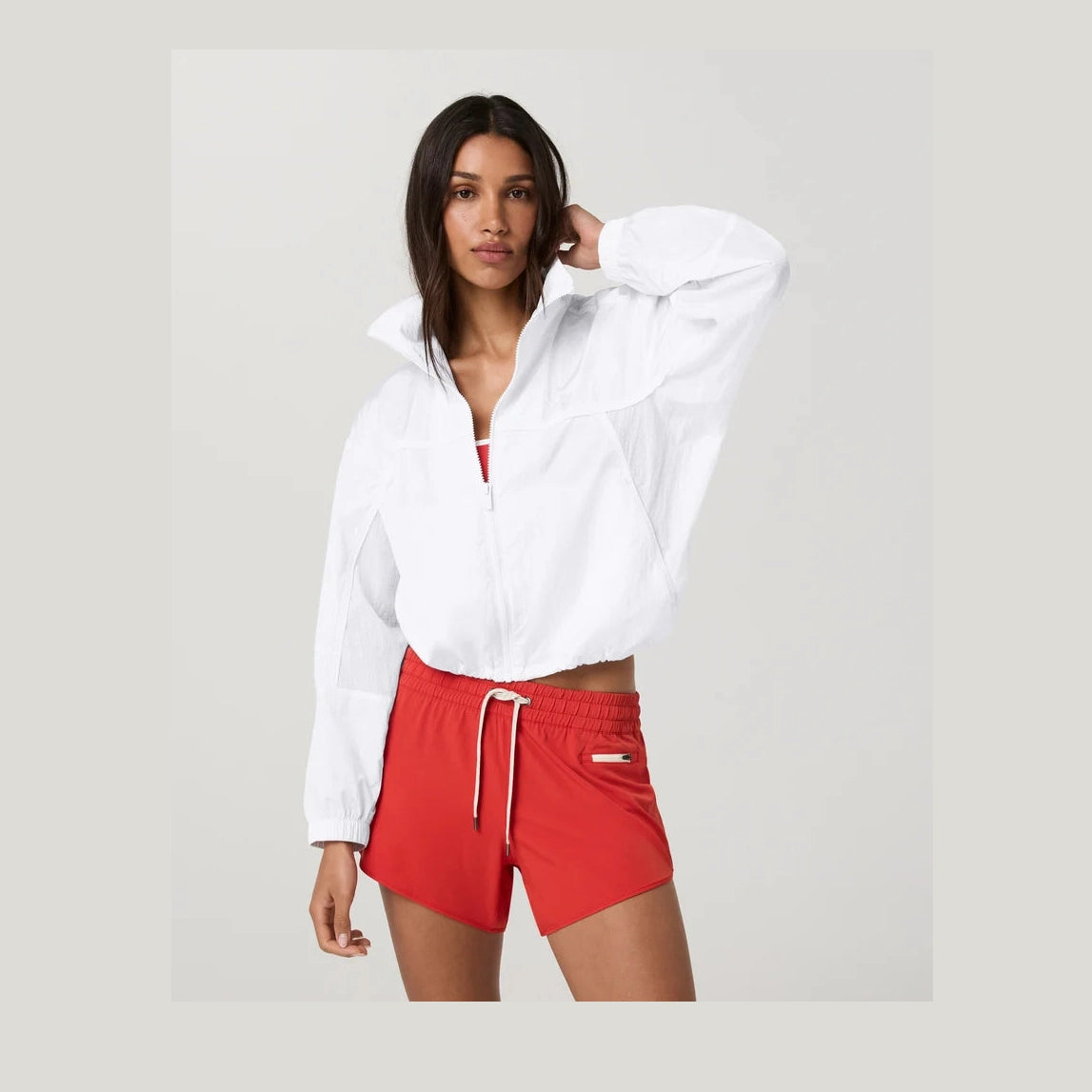 Woman wearing a white Court Full Zip--Women's jacket and red shorts on a plain background