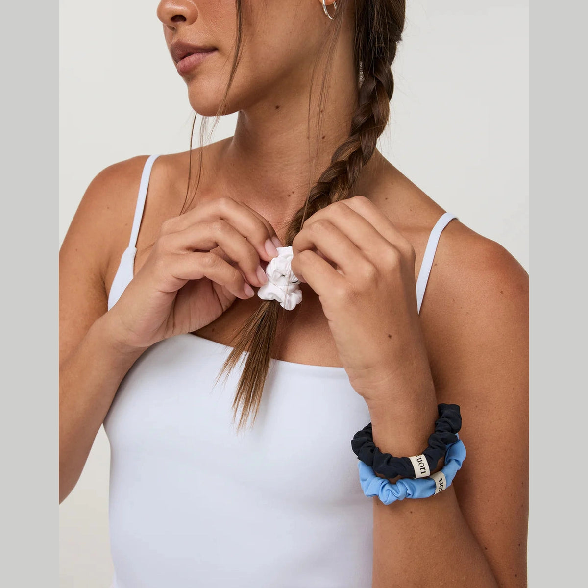 A close-up of a woman with a braided ponytail adjusting a white scrunchie, wearing a stack of three scrunchies in navy, light blue, and white on her wrist against a plain light background.