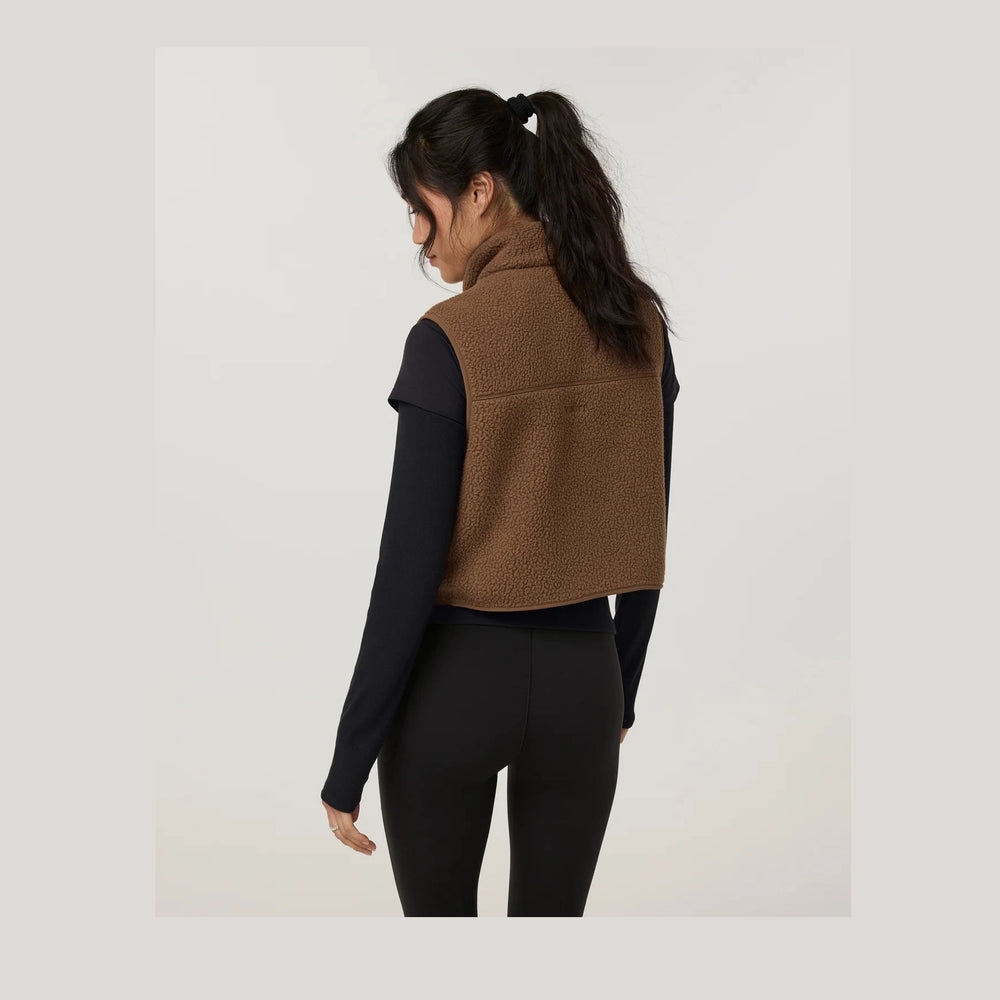 A woman shown from the back wearing a brown sherpa fleece cropped vest over a black long-sleeve top and black leggings, against a plain light gray studio background.