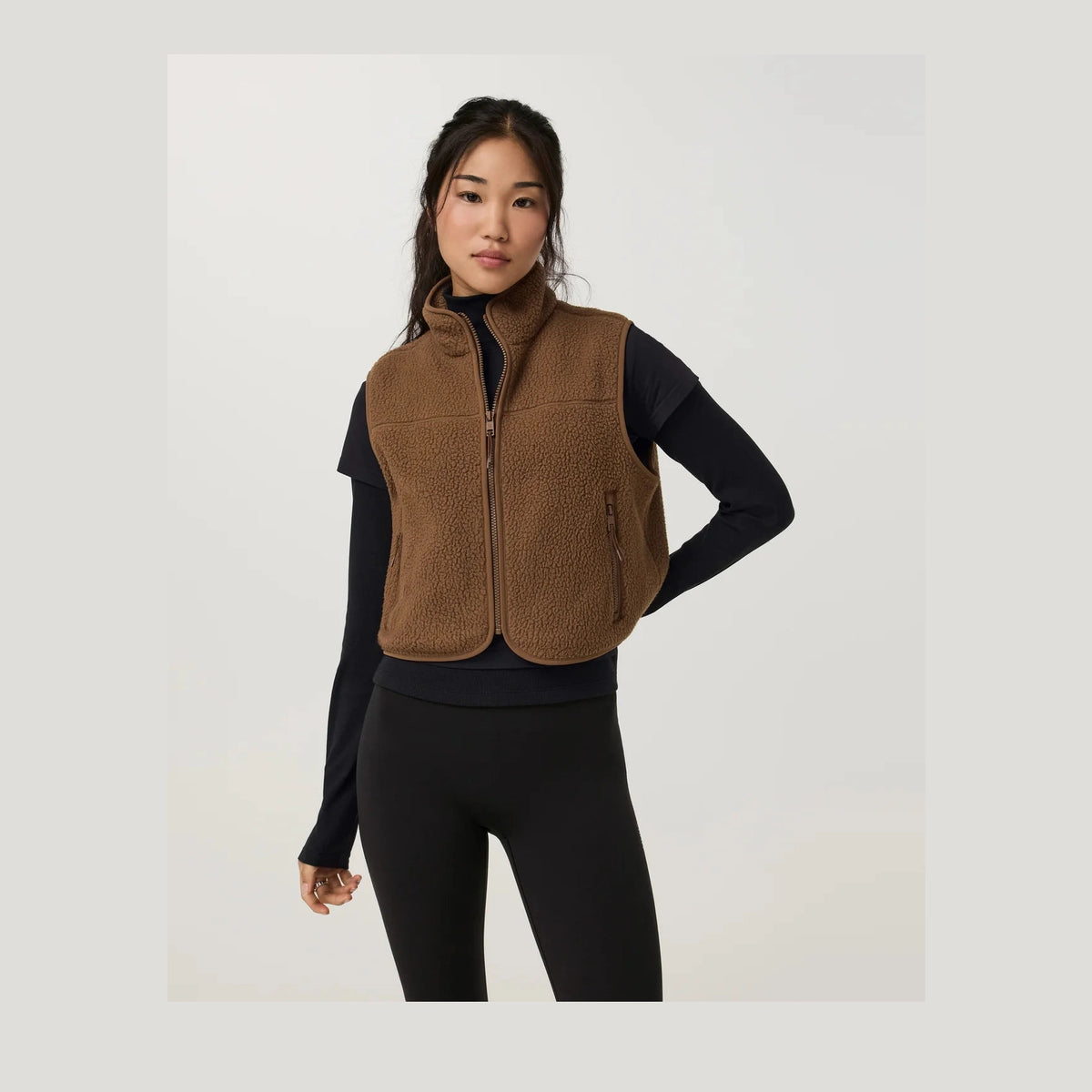 A woman modeling a brown fleece Alpine Vest--Women's over a black long-sleeve top and black leggings, photographed against a clean light gray studio background.