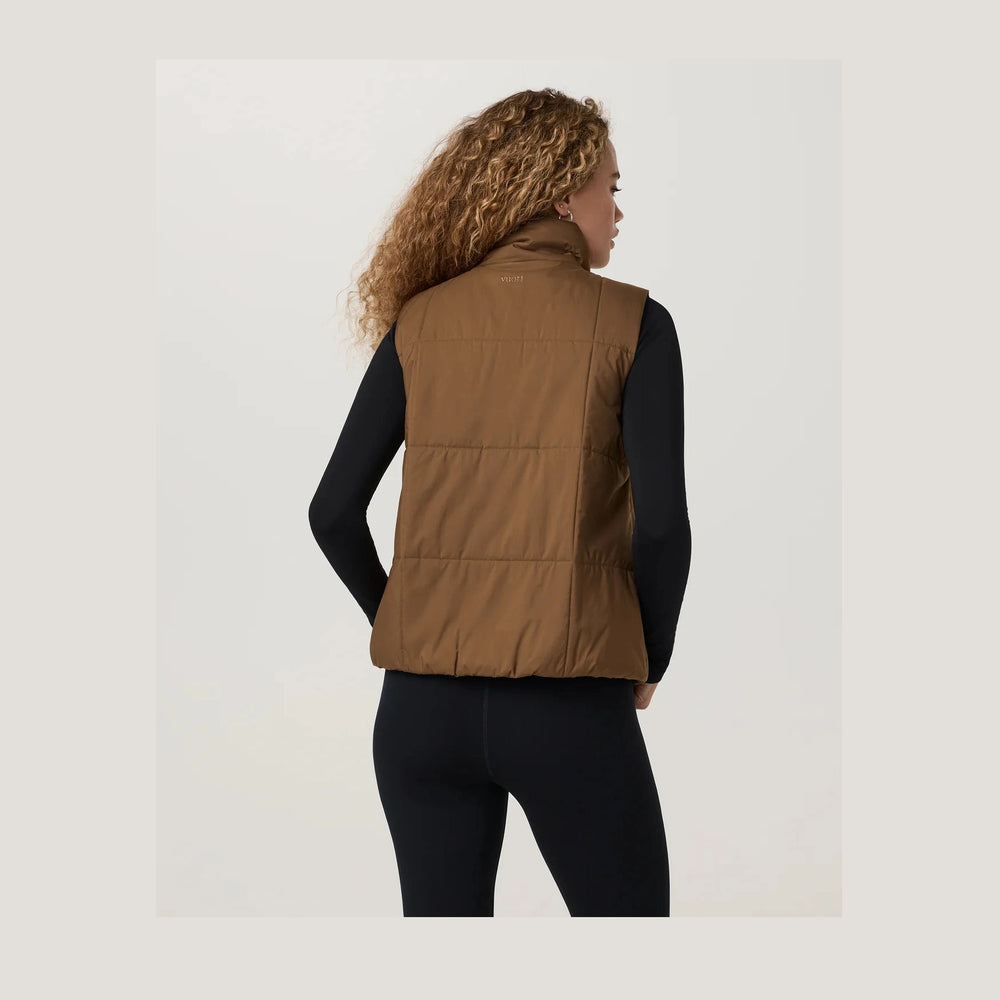 A woman shown from the back wearing a brown Canyon Insulated Sporty Vest--Women's over a black long-sleeve top and black leggings, photographed against a clean light gray studio background.