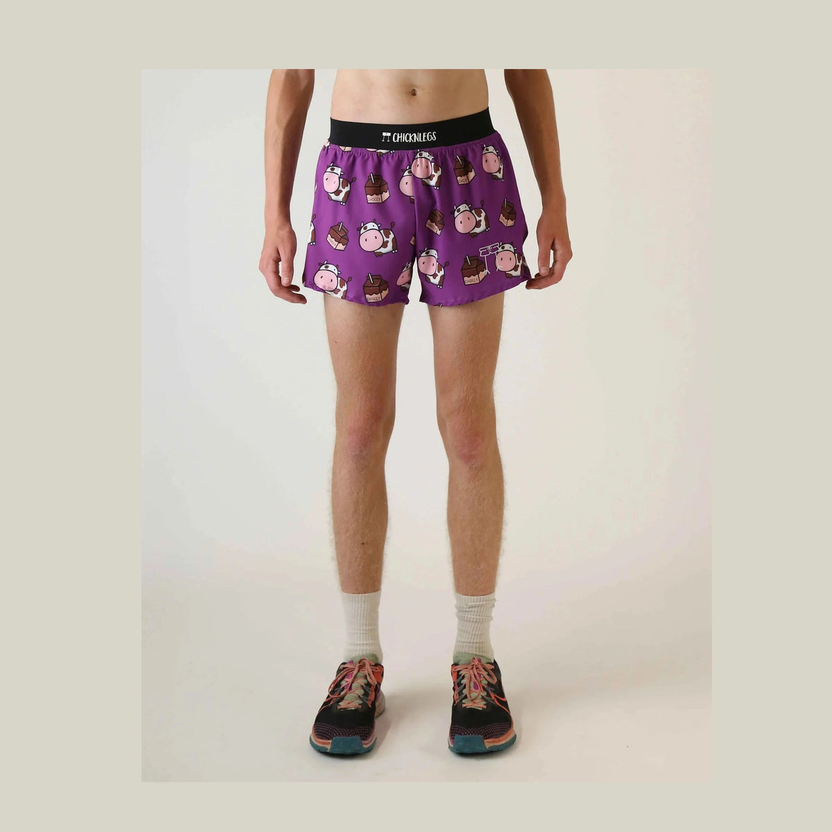 A model is shown from the waist down wearing purple Chicknlegs 4" Half Split Shorts – Men's with a cartoon cow print, a black waistband with the brand name 'Chicknlegs', white socks, and running shoes against a plain light background.