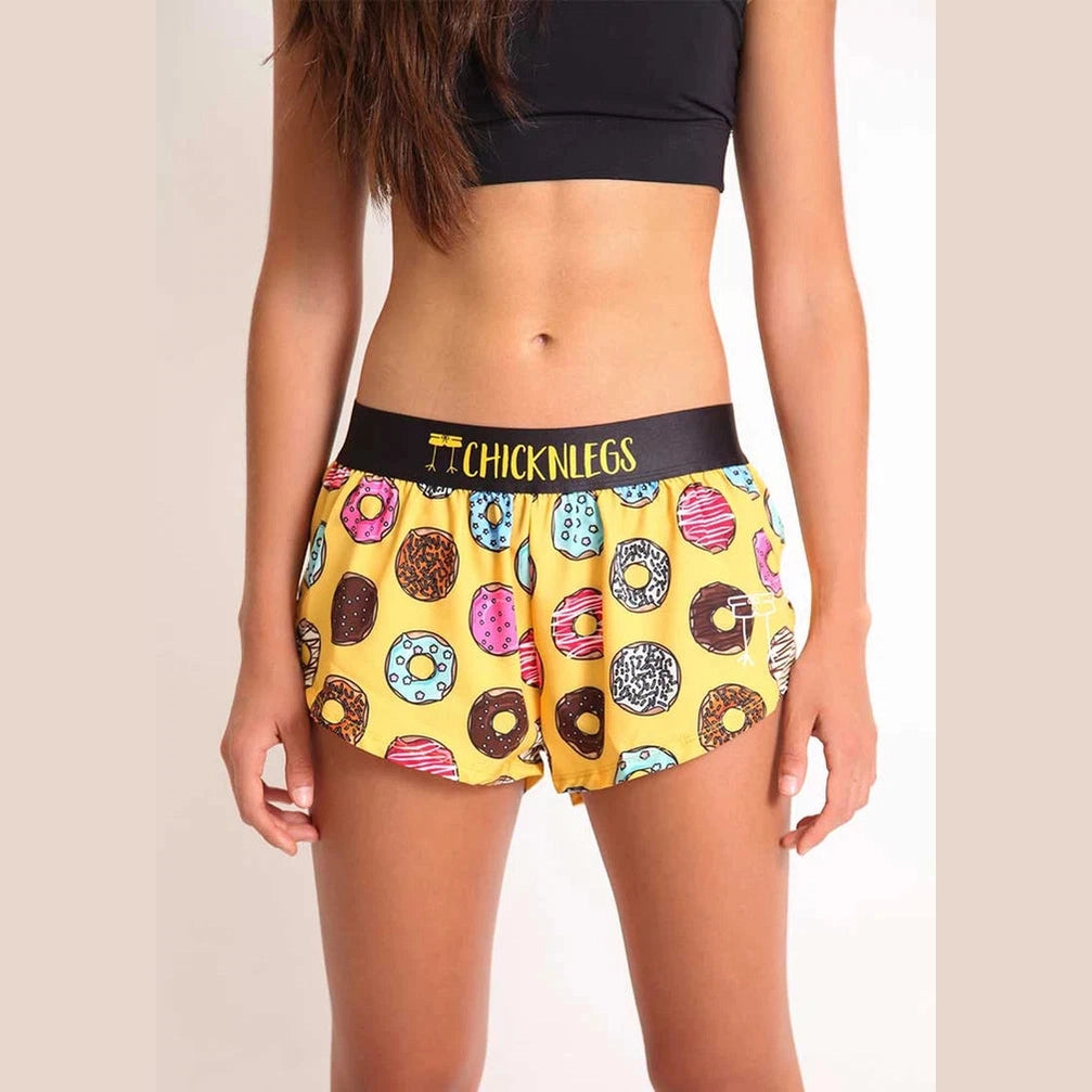 A model wearing yellow Chicknlegs 1.5" Split Shorts – Women's with a colorful donut print and a black waistband featuring the brand name 'CHICKNLEGS' in gold lettering.