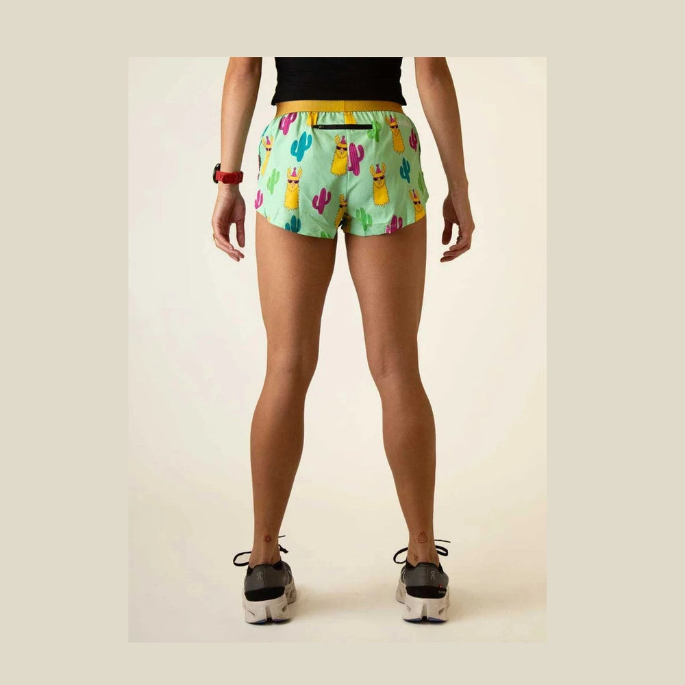 Rear view of a model wearing light green Chicknlegs 1.5" Split Shorts – Women's with a colorful llama and cactus print, paired with a black top and black sneakers, against a plain beige background.