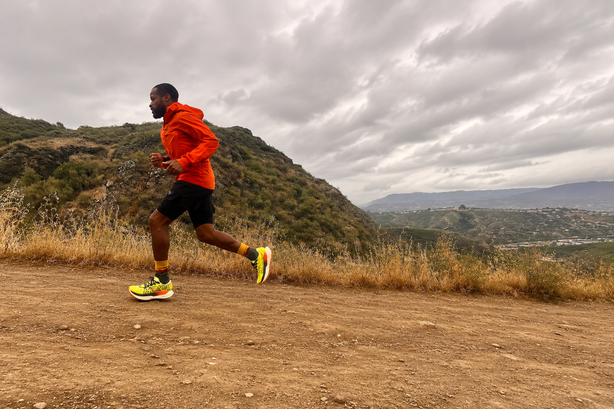 A male runner in an orange hooded jacket, black shorts, and bright yellow running shoes is jogging on a dirt trail with a mountain landscape and cloudy sky in the background.