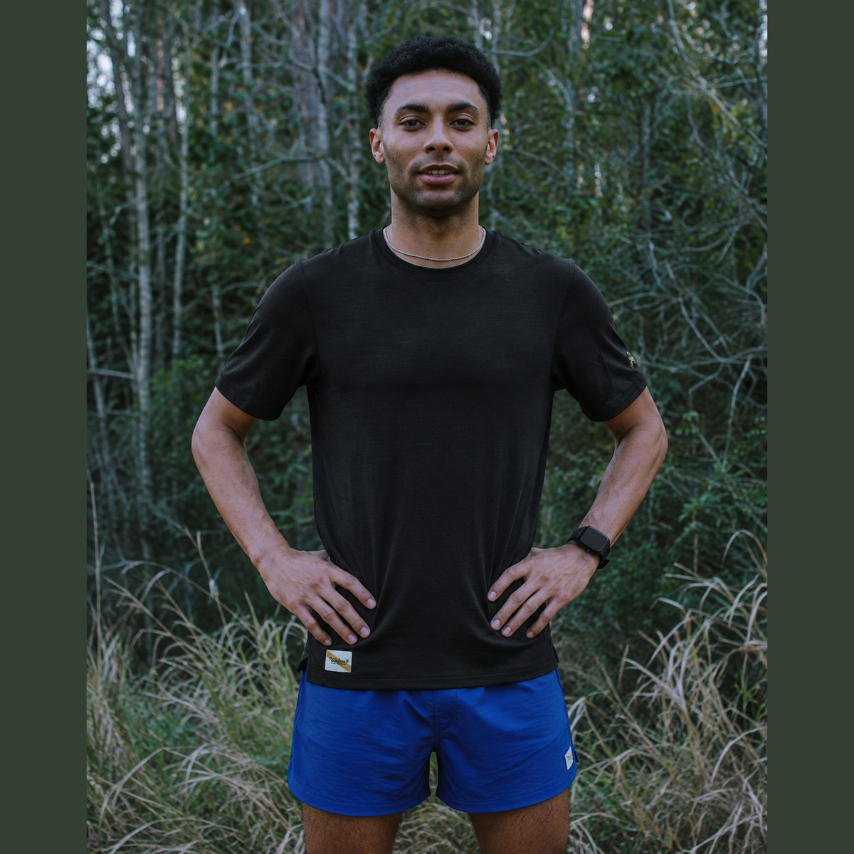 A man wearing a black Session Tee – Men's athletic t-shirt and blue running shorts stands outdoors in a grassy, wooded area with trees in the background.