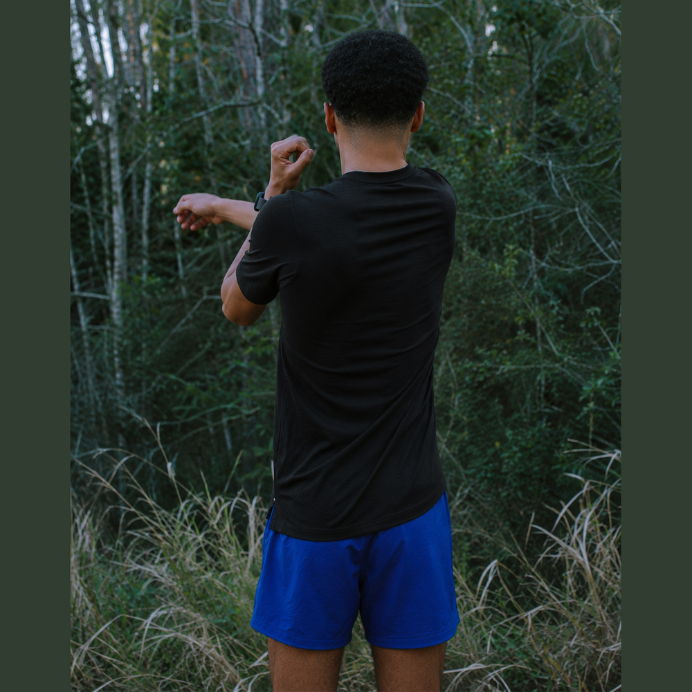 A man viewed from behind wearing a black Session Tee – Men's athletic t-shirt and bright blue running shorts, standing in a grassy outdoor forest setting.