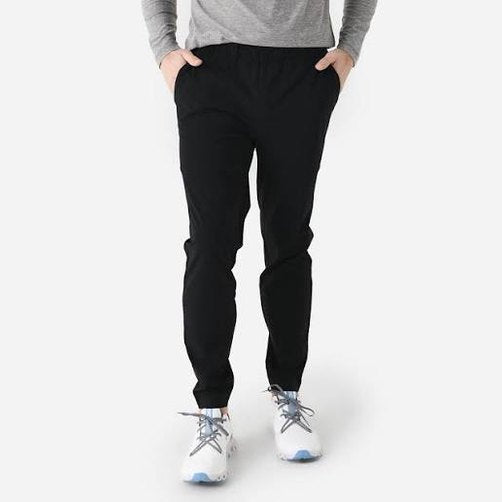 A person wearing black Vuori Fleet Pant – Men's pants, a gray long-sleeve top, and white sneakers, standing against a light gray studio background.
