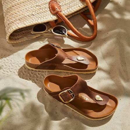 A pair of brown leather thong sandals with gold-tone buckle details, styled on a sandy surface next to a woven straw bag and sunglasses.