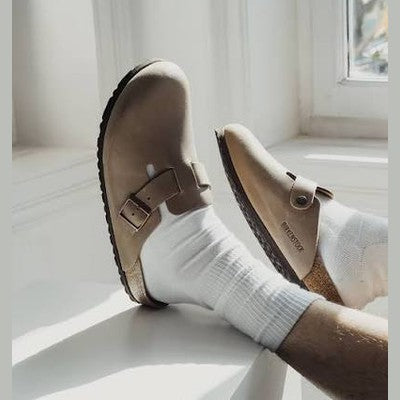 A person wearing white crew socks and tan Birkenstock Bungee clogs, sitting by a bright window.