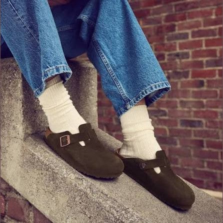 A person sitting on concrete steps wearing blue jeans, white socks, and dark olive-brown clog-style mules with metal buckle straps. A red brick wall is visible in the background.