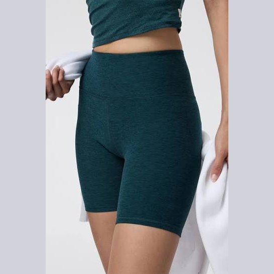 A close-up of a model wearing high-waisted dark teal heather Vuori Clean Elevation Shorty – Women's biker shorts, holding a white towel against a light gray studio background.