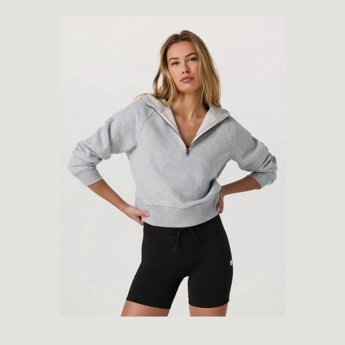 A woman modeling a light gray heathered Vuori Restore Half Zip Hoodie – Women's with a contrasting white inner collar, paired with black bike shorts, against a plain white studio background.