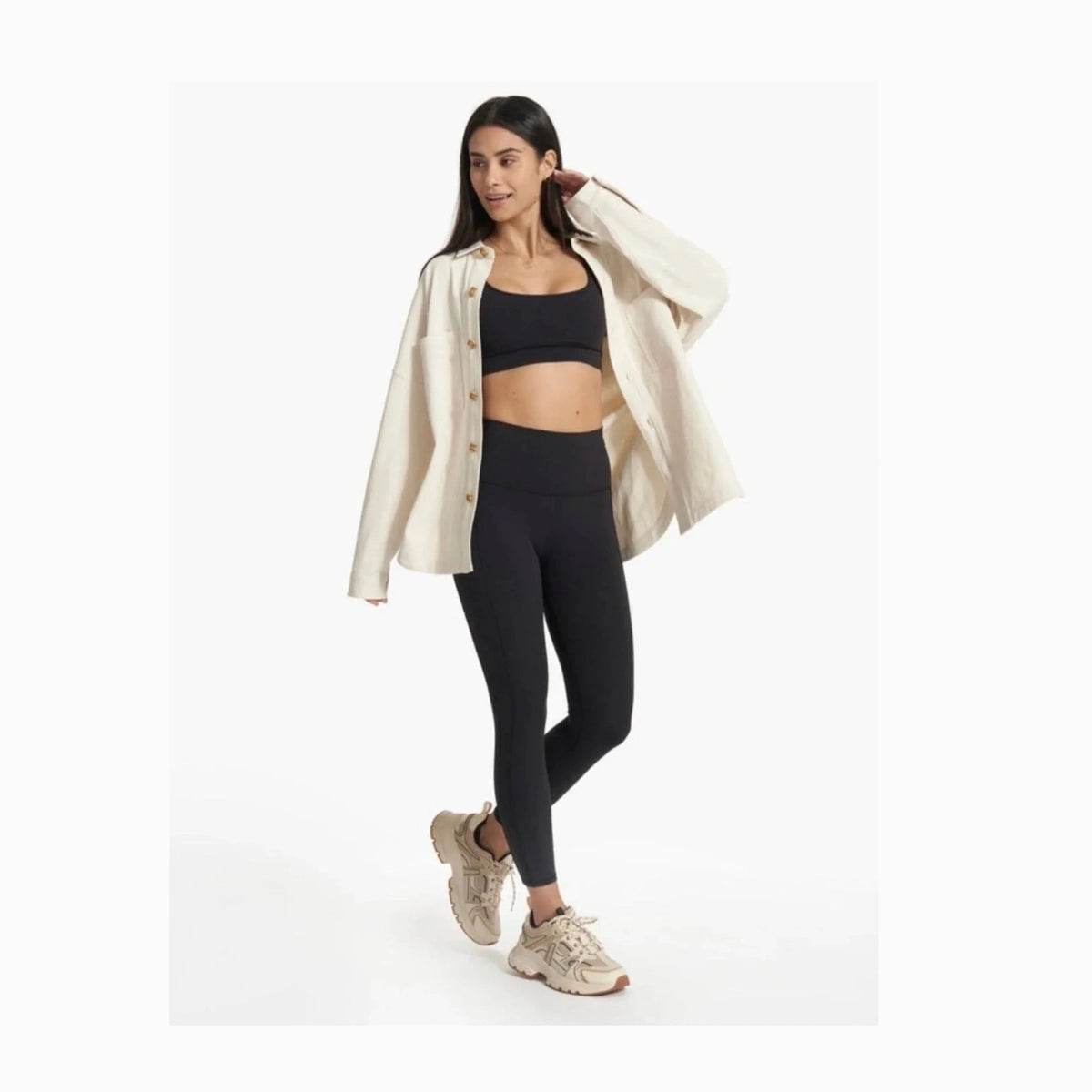 A woman modeling a black sports bra and high-waisted black leggings, styled with an open cream-colored button-up Vuori Mackenzie Shirt Jacket – Women's shirt and beige sneakers, against a plain white studio background.
