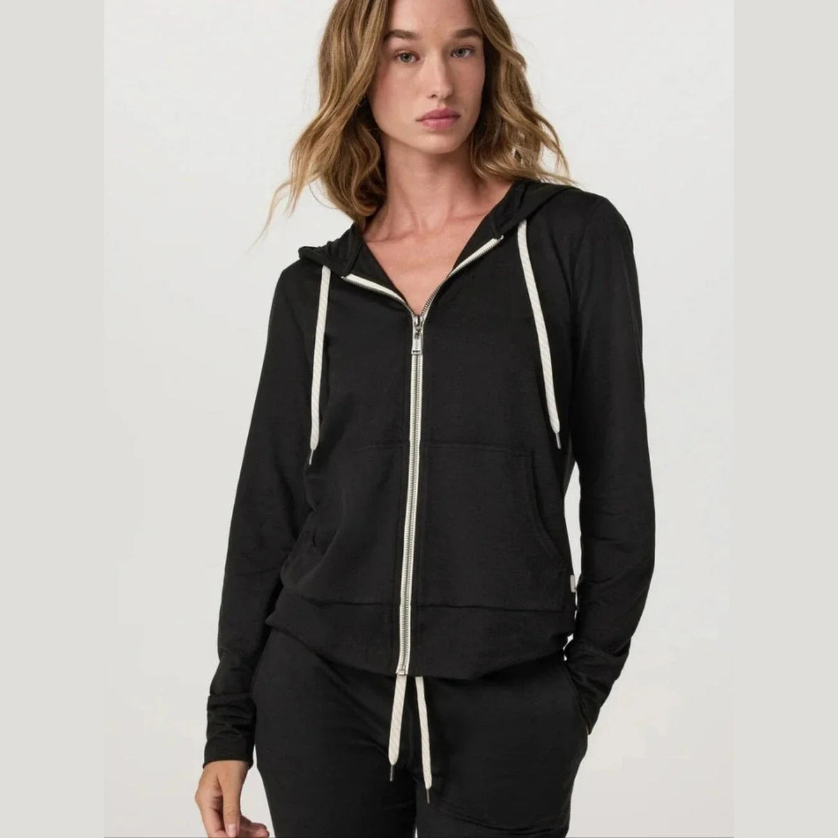 A woman modeling a black Vuori Halo Performance Full Zip Hoodie – Women's hoodie with white drawstrings and a white front zipper, paired with black pants, against a light gray studio background.