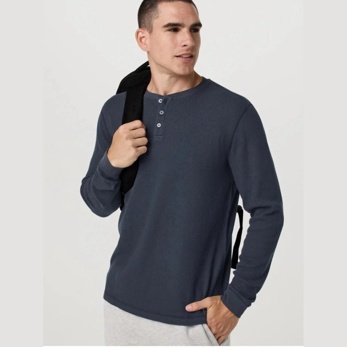 A man wearing a dark slate blue Vuori Waffle Henley – Men's long-sleeve henley shirt, holding a black backpack strap over his shoulder, against a plain light gray background.
