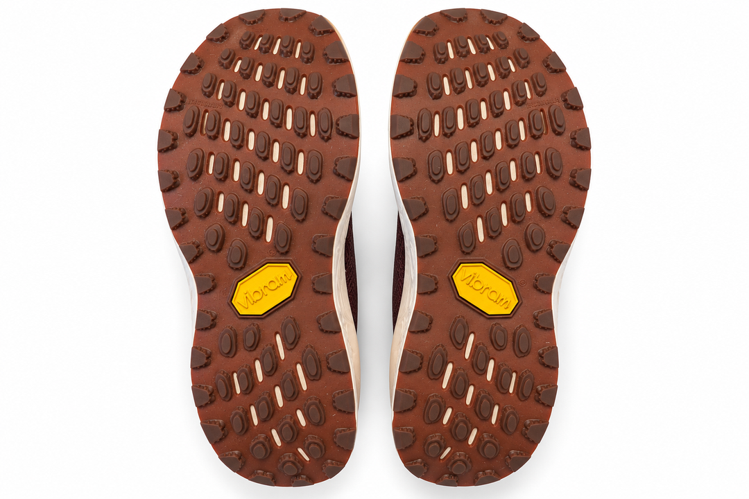 Top-down view of a pair of brown Vibram hiking shoe soles, showing the tread pattern and the signature yellow Vibram logo on a white background.