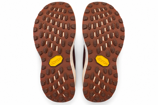 Top-down view of a pair of brown Vibram hiking shoe soles, showing the tread pattern and the signature yellow Vibram logo on a white background.