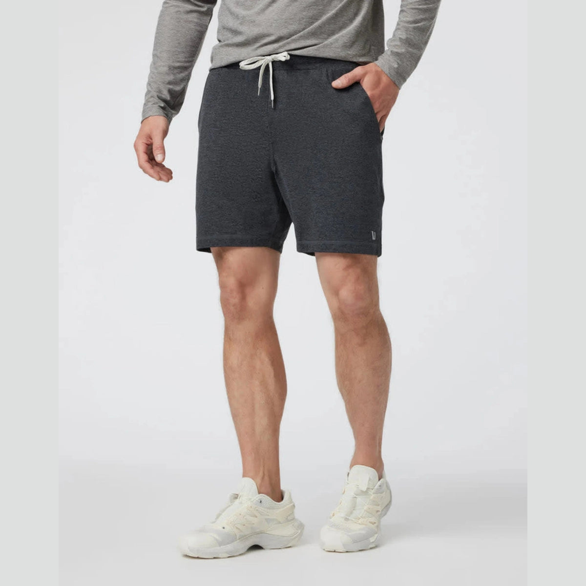 A man wearing dark heather gray Vuori Ponto Short – Men's  athletic shorts with a white drawstring, paired with a gray long-sleeve shirt and white sneakers, standing against a light gray studio background.