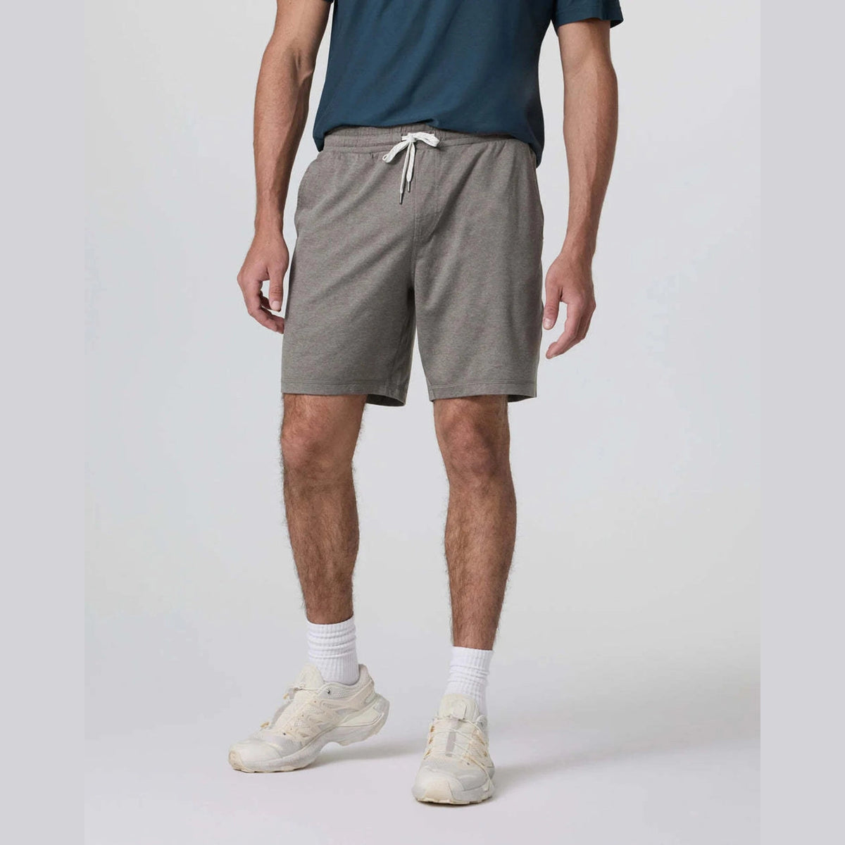 A man wearing heather gray Vuori Ponto Short – Men's drawstring shorts, a dark teal t-shirt, white socks, and off-white sneakers, standing against a light gray studio background.